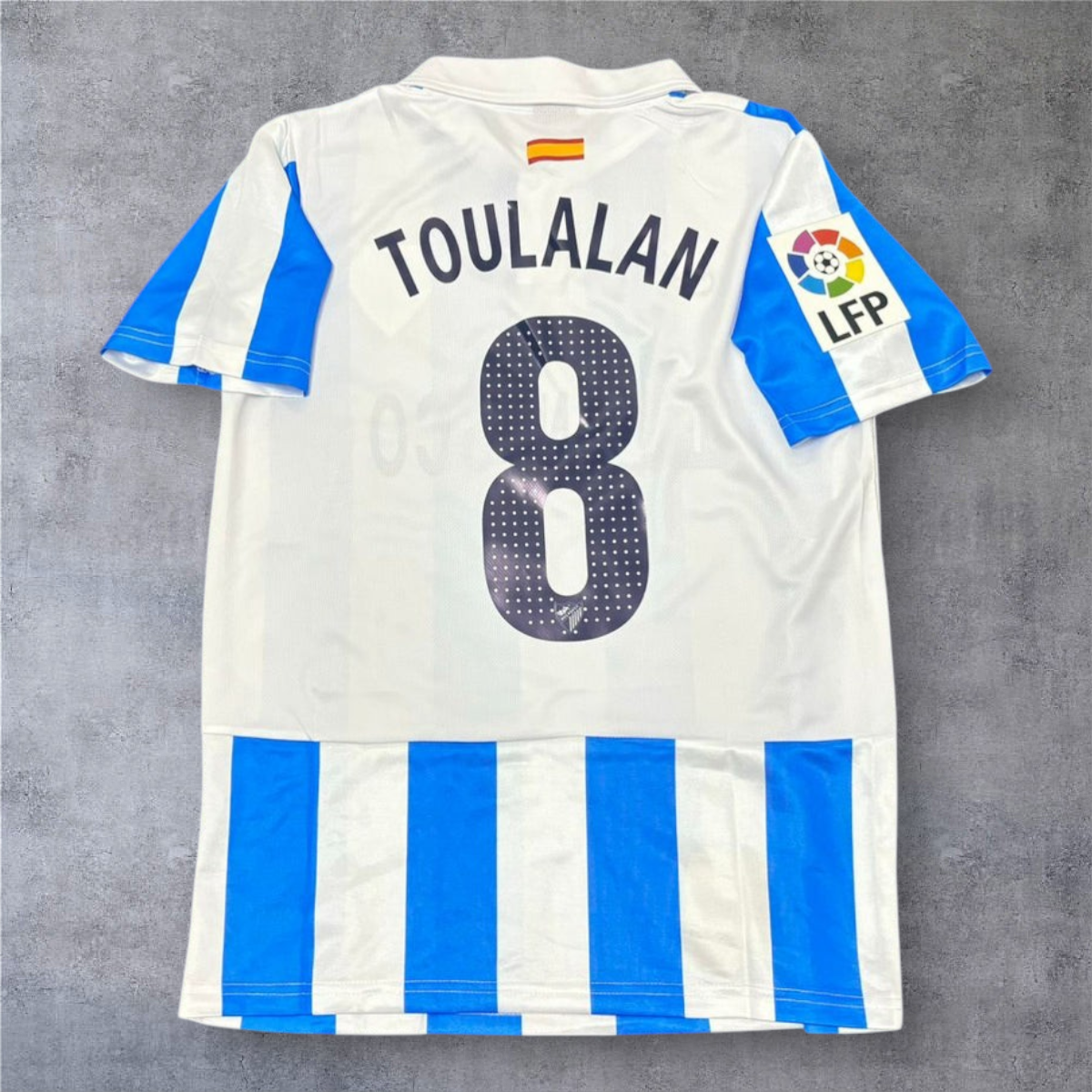 Camisola de futebol A minha loja Málaga 2012-13, branca e azul, com nome Toulalan e número 8 nas costas.