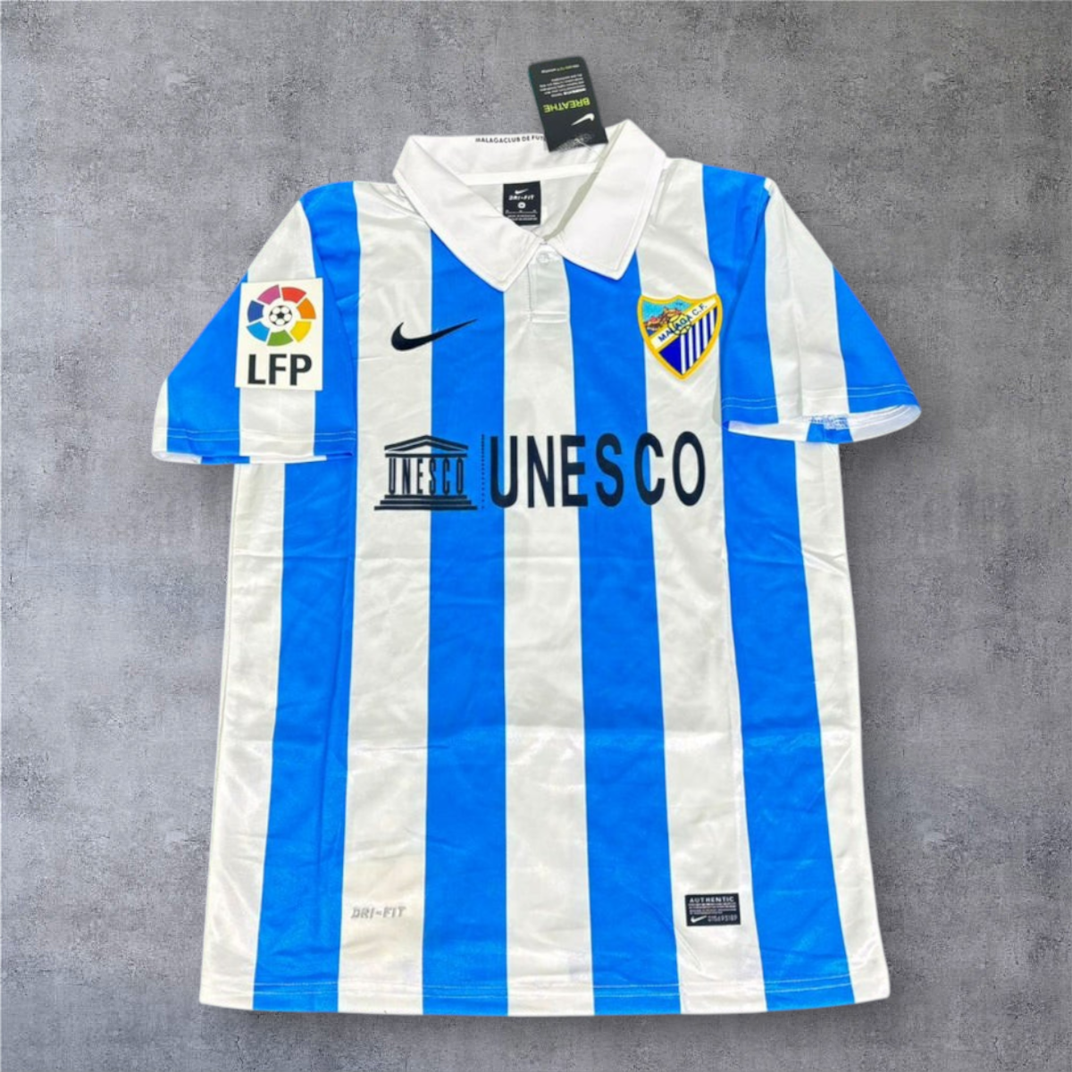 Camisola de futebol A minha loja Málaga 2012-13, listada em azul e branco com gola polo e patrocinador UNESCO.