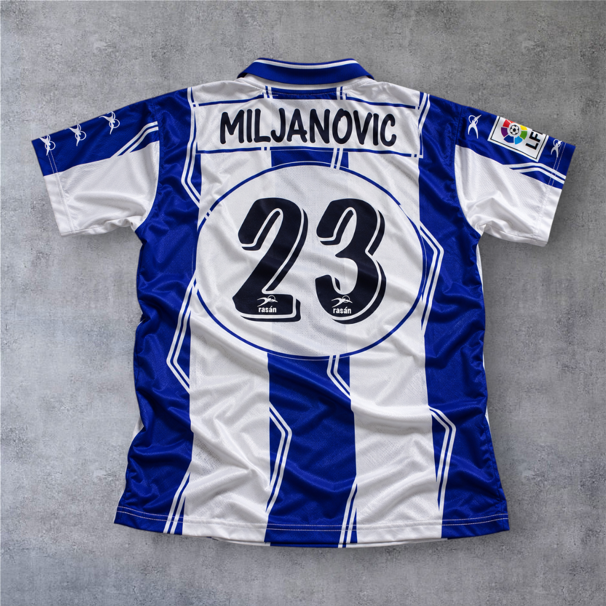 Camisola de futebol A minha loja Hercules 1997/98, listada em azul e branco com o nome Miljanovic e número 23 atrás.